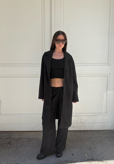 Abrigo largo negro sobre un top corto negro ajustado y pantalones sueltos negros, combinados con botas negras gruesas y grandes gafas de sol negras.