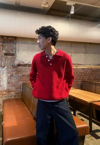 Jeune homme en pull en laine rouge et jean foncé, debout, les mains derri ère le dos, dans un café aux murs en briques et aux banquettes marron.