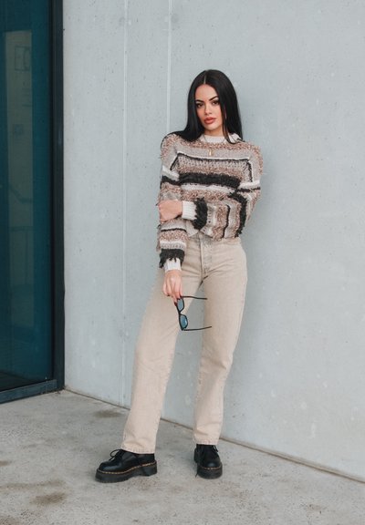 Suéter de punto texturizado en rayas beige, marrón y negro; jeans de tiro alto color crema; zapatos de plataforma negros; sosteniendo unas gafas de sol.