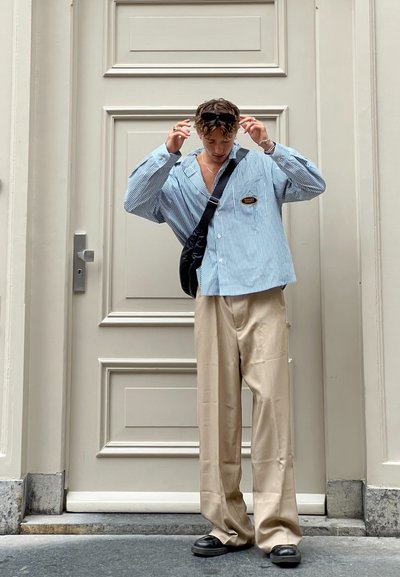 Camisa oversized a rayas azules, pantalones de pierna ancha en beige, gafas de sol negras y un bolso cruzado negro, de pie contra una puerta neutral.