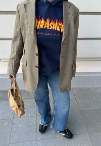 Υφασμάτινο καφέ σακάκι houndstooth πάνω από ένα σκούρο μπλε hoodie με μια φλογερή λογότυπο; ανοιχτό μπλε τζιν; μαύρα παπούτσια; κρατώντας μια μπεζ τσάντα.