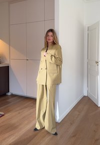 Blazer jaune clair et pantalon large assorti, en tissu tissé. Le blazer est équipé de deux boutons et de poches plaquées.
