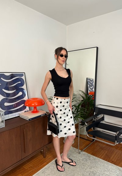 Mujer con camiseta sin mangas negra y falda blanca con lunares sosteniendo bolso negro, de pie en una habitación moderna con espejo y lámpara naranja.