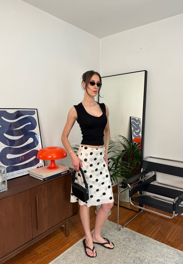 Frau in schwarzem Tanktop und weißem gepunktetem Rock hält eine schwarze Handtasche und steht in einem modernen Zimmer mit Spiegel und orangefarbener Lampe.