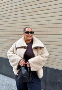 Naine kannab üle suurusega kreemikat taljesoengu jopet, tumedaid päikeseprille ja kuldseid ehteid, hoides musta käekotti beeži tellismüüri taustal.