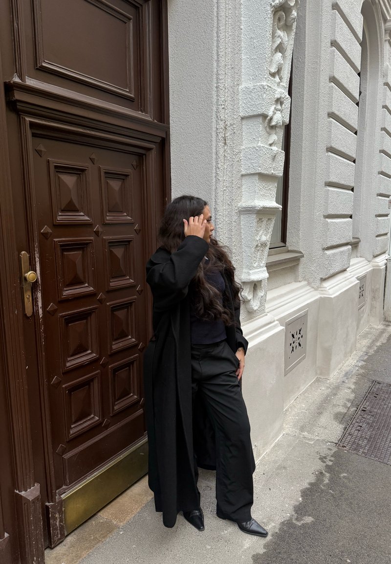 Longue veste noire et pantalon noir ample portés sur un haut sombre. La porte est brun foncé avec un panneau géométrique, adjacente à une architecture blanche texturée.