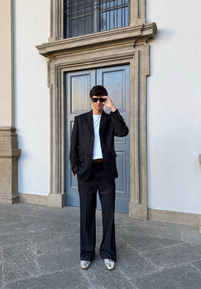 Blazer negro con rayas finas a juego con pantalones acampanados anchos, combinado con una camiseta blanca y zapatillas plateadas, frente a una puerta de color azul claro.