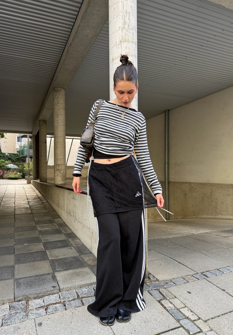 Joven mujer vistiendo una camiseta de manga larga a rayas, pantalones negros anchos con rayas blancas y una falda negra superpuesta, de pie junto a una columna al aire libre.