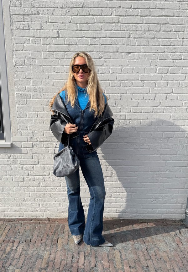 Nő hosszú szőke hajjal, napszemüveget visel, kék garbó, farmer overál, fekete bőrdzseki, szürke kézitáskát tart, fehér téglafal előtt áll.