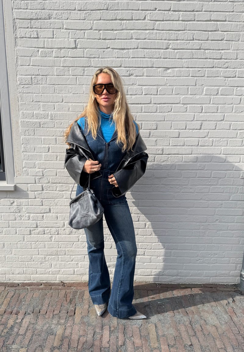 Γυναίκα με μακριά ξανθά μαλλιά, που φοράει γυαλιά ηλίου, μπλε ζιβάγκο, τζιν ολόσωμη φόρμα, μαύρο δερμάτινο μπουφάν, κρατώντας γκρι τσάντα, στέκεται μπροστά σε λευκό τοίχο από τούβλα.