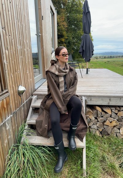 Abrigo largo marrón sobre suéter gris, mallas negras y botas de goma negras. Los accesorios incluyen una bufanda estampada y gafas de sol. Fondo de madera.