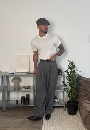 Hombre con tatuajes vestido con camiseta blanca, pantalones anchos grises y gorra plana gris, de pie en el interior cerca de estanterías con decoración y una planta en maceta.