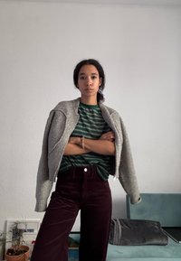 Lys grå strikket jakke over en grøn- og sortstribet skjorte, parret med højtsiddende bordeaux fløjlshavnerbukser. Minimalistisk baggrund.