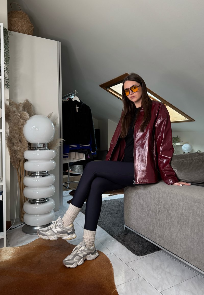 Femme portant des lunettes de soleil orange, une veste en cuir rouge, des leggings noirs et des baskets assise sur un canapé gris dans une pièce mansardée moderne.