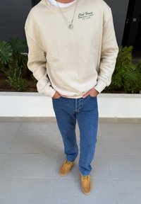 Kremowy sweter z logo, zestawiony z niebieskimi dżinsami i jasnobrązowymi butami z żółtymi sznurowadłami. Prosty design, casualowy krój.