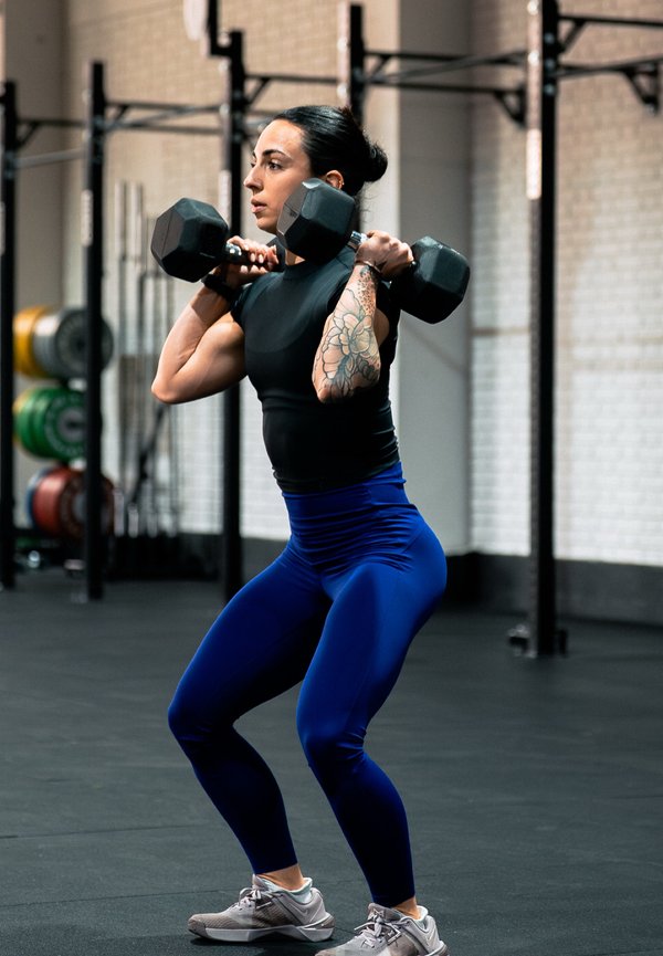 Woman with tattooed arm lifting two dumbbells at shoulder height in a gym, preparing for a squat exercise.