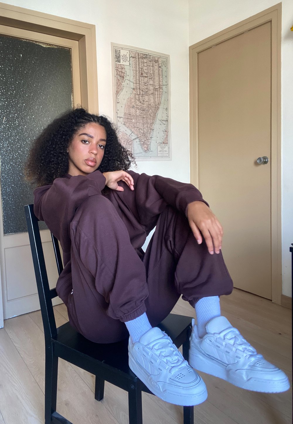 Sudadera marrón de gran tamaño y joggers a juego, zapatillas blancas y calcetines azul claro. Cabello rizado, sentado en una silla negra frente a una pared clara.