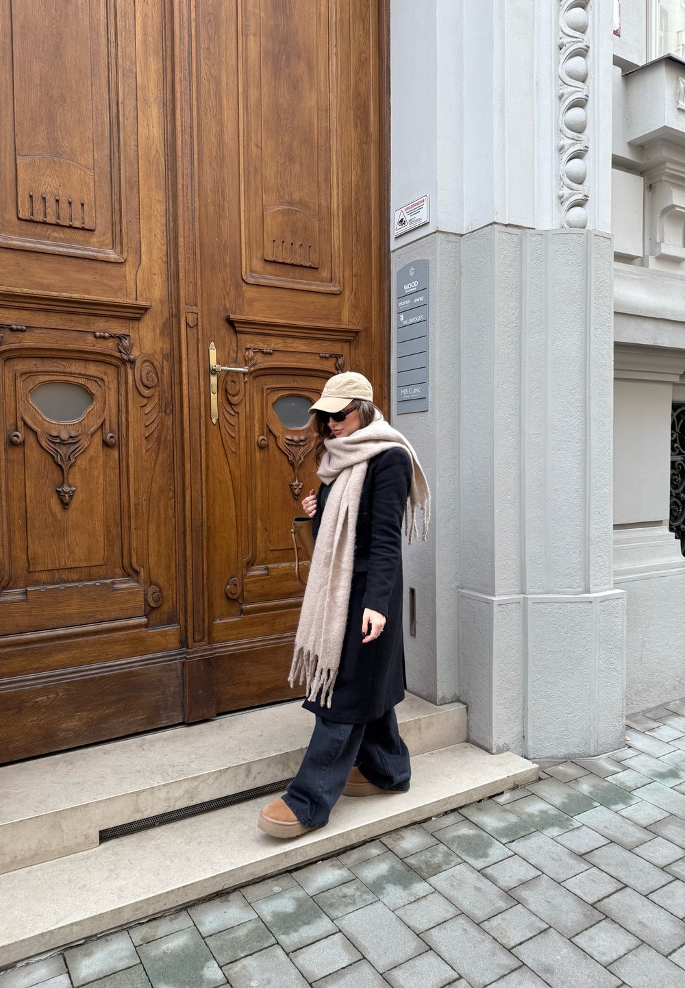 Μαύρο παλτό, μπεζ κασκόλ, καπέλο και καφέ μποτάκια. Ξύλινες διπλές πόρτες με περίπλοκα σχέδια και ελαφριά γκρίζα πρόσοψη κτιρίου. Επίστρωση δρόμου.