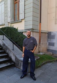 Mężczyzna w okularach i tatuażu, ubrany w pasiastą koszulę i szerokie dżinsy, stoi obok kamiennych schodów i narożnika budynku na zewnątrz.