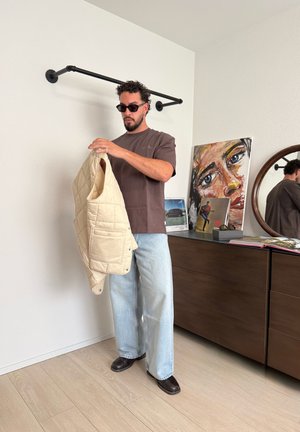 Hombre con gafas de sol sostiene un chaleco beige, de pie junto a una cómoda con arte, discos y un espejo redondo en una habitación llena de luz.