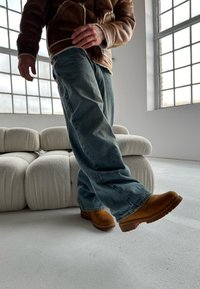 Giacca di pelle marrone, jeans di denim blu e stivali beige slip-on. La stanza presenta grandi finestre e un divano bianco dalla texture particolare.