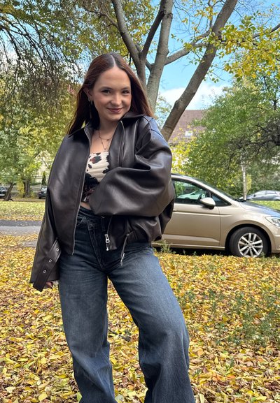 Joven mujer con chaqueta de cuero negra y jeans sonríe de pie en un suelo cubierto de hojas, con árboles y coches aparcados al fondo.