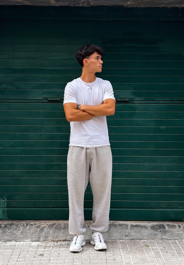Jonge man in een wit T-shirt en grijze joggingbroek staat met armen over elkaar, kijkt naar de zijkant tegen een donkergroene metalen deur.