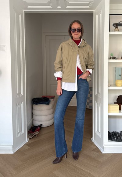 Mujer con chaqueta beige, camisa blanca, cuello alto rojo, vaqueros azules y zapatos de tacón marrones, de pie en el interior cerca de una puerta y una estantería.