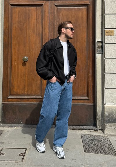 Hombre que lleva una chaqueta negra, camisa blanca, vaqueros azules, gafas de sol y zapatillas blancas, de pie con las manos en los bolsillos frente a una gran puerta de madera.