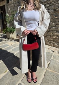 Sac en cuir rouge avec une forme incurvée et une courte anse, tenu par une personne portant un manteau beige et un pantalon noir, debout sur un pavé en pierre.