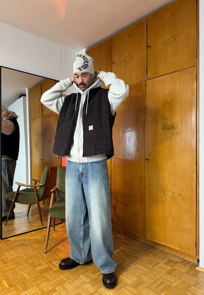 Hombre con sudadera con capucha gris, chaleco negro, jeans anchos azules y gorro blanco ajustándose la capucha en una habitación con armarios de madera y una silla verde.
