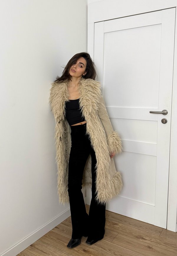 Femme appuyée contre un mur portant un manteau beige long bordé de fourrure, un crop top noir, un pantalon noir évasé et des chaussures pointues noires.