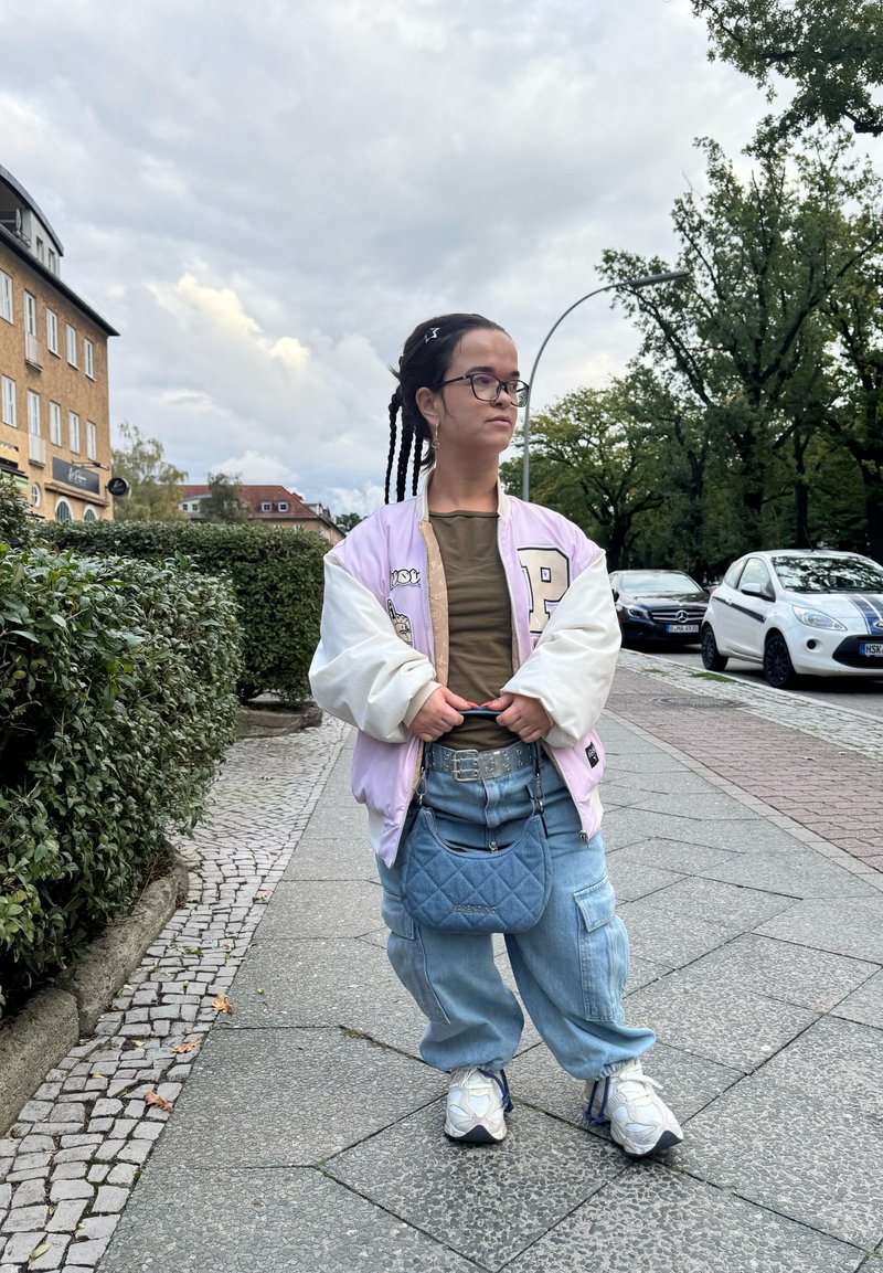 Ροζ και λευκή bomber jacket, ελιάς πράσινη μπλούζα, φαρδιά μπλε τζιν παντελόνια, καπιτονέ μπλε τσάντα και λευκά αθλητικά παπούτσια.