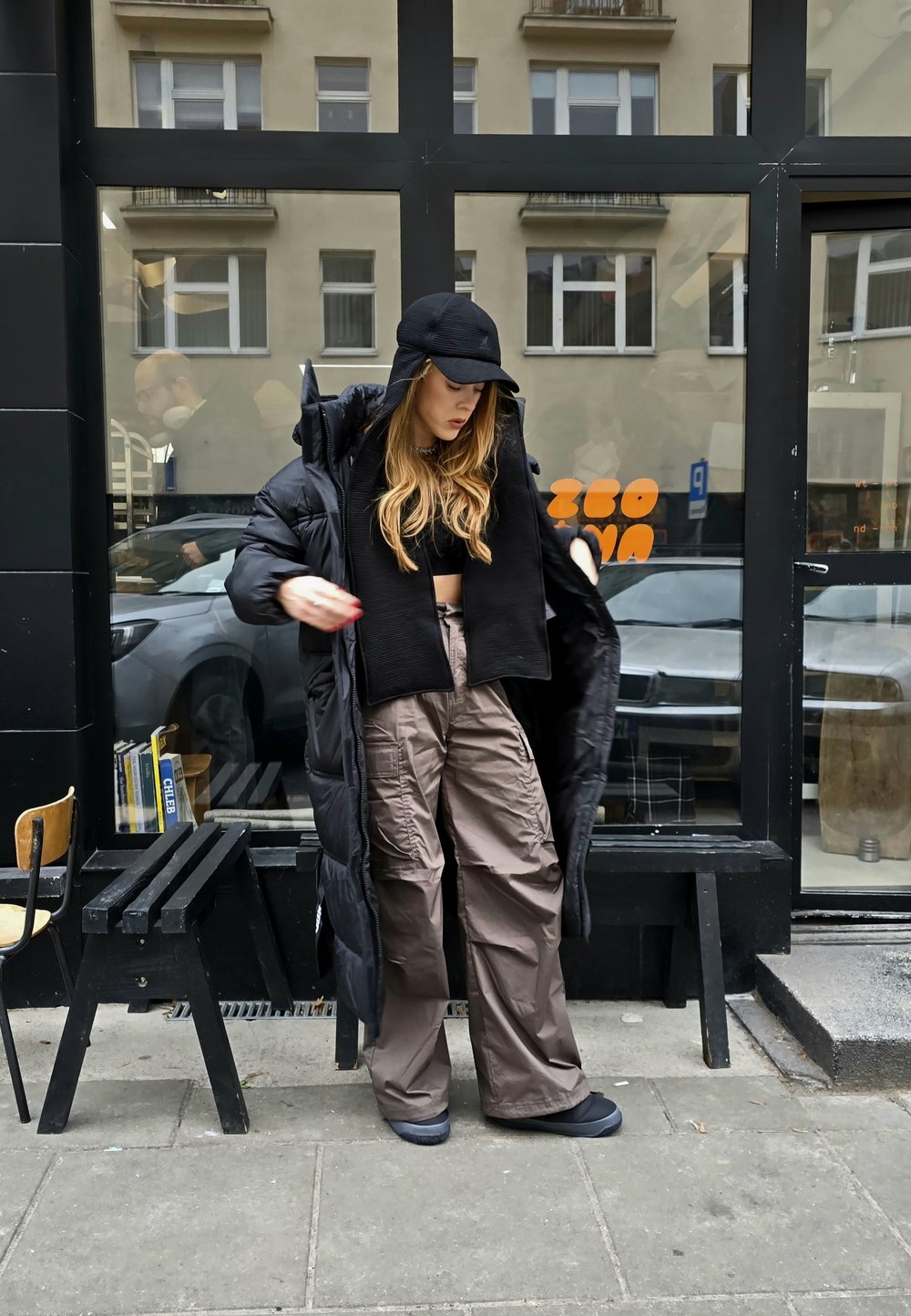 Sort pufferjakke båret åben over en sort crop top, med brune cargo bukser og sorte sko. Inkluderer en kasket og langt, bølget hår.