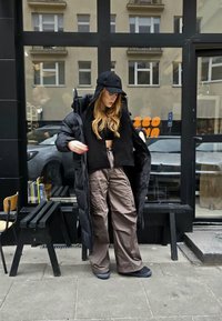 Sort pufferjakke båret åben over en sort crop top, med brune cargo bukser og sorte sko. Inkluderer en kasket og langt, bølget hår.