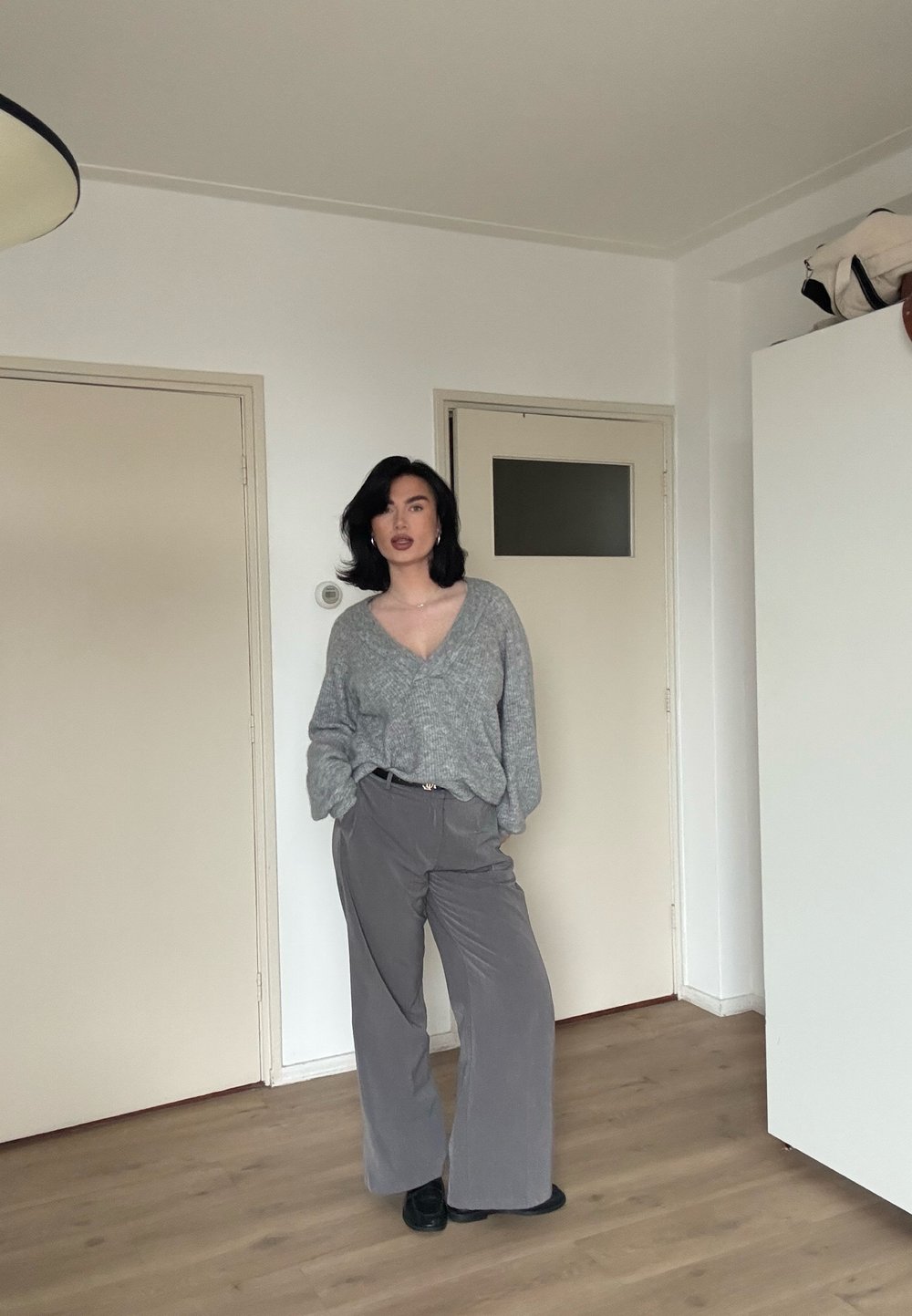 Pull en maille grise avec un col en V, associé à un pantalon gris à jambes larges. Texture lisse et coupe décontractée ; debout dans une pièce simple.