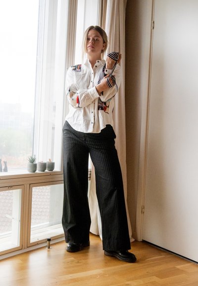 Camisa blanca de botones con acentos gráficos coloridos, combinada con pantalones de pierna ancha de rayas negras. De pie junto a una ventana con plantas cercanas.