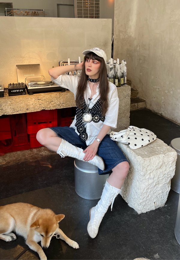 Jeune femme en bottes blanches et casquette assise sur un tabouret métallique près d'un bloc de pierre avec un sac à pois, tandis qu'un chien est couché sur le sol dans une pièce avec du matériel de DJ.