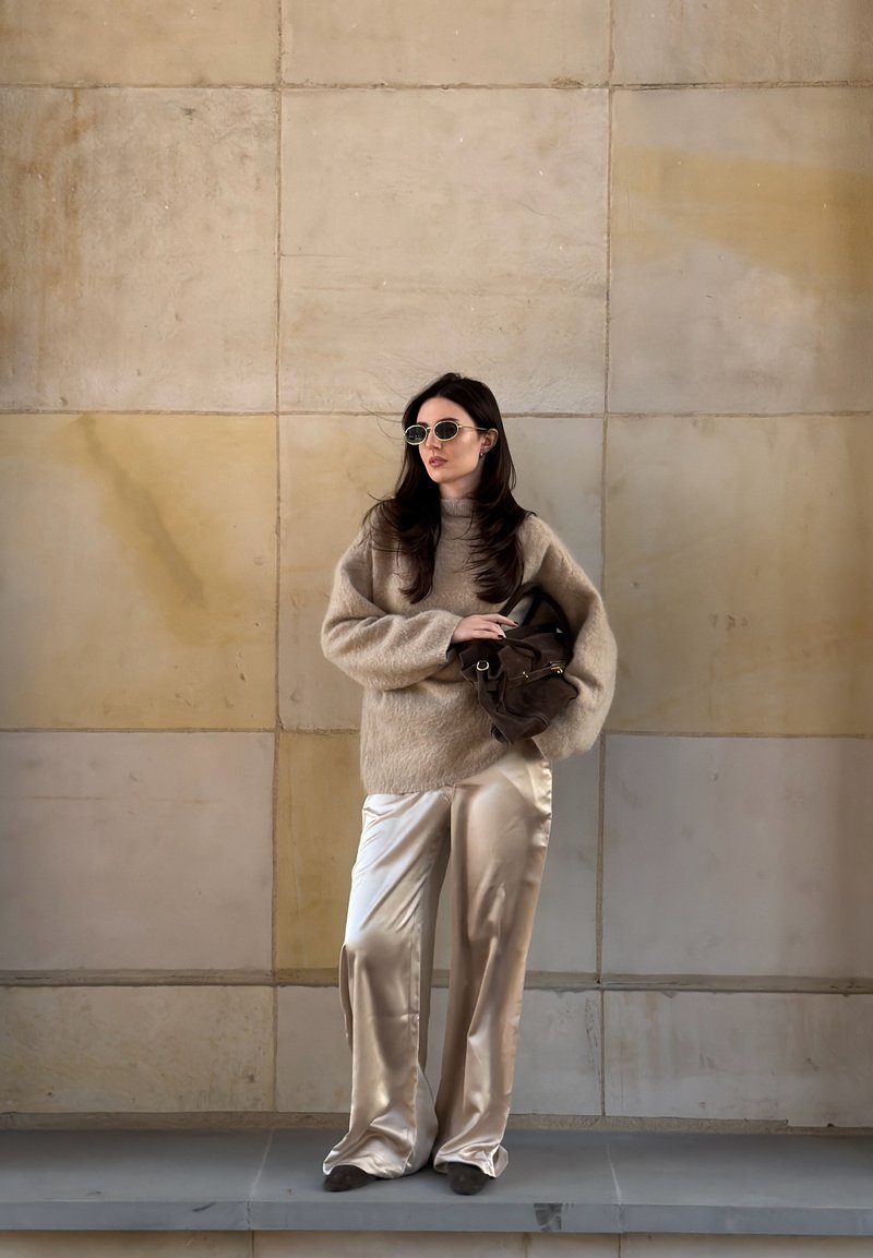 Donna con maglione beige peloso, pantaloni larghi di raso, occhiali da sole tondi, che tiene una borsa marrone contro una parete piastrellata color sabbia.