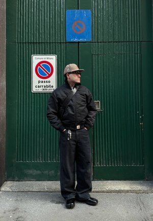 Hombre con chaqueta negra, pantalones anchos negros y gorra de cuadros, de pie con las manos en los bolsillos frente a una puerta verde con señales de no aparcar.