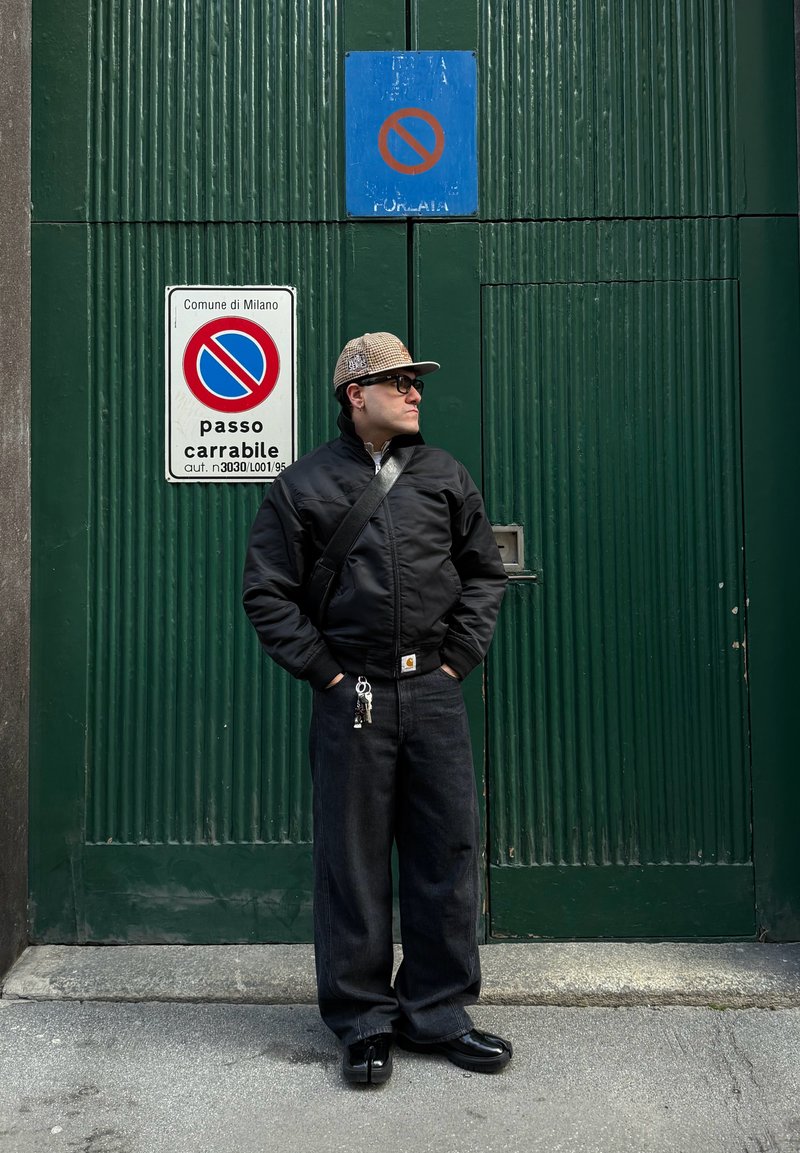 Man in black jacket, wide black trousers, and checkered cap stands with hands in pockets in front of a green door with no parking signs.