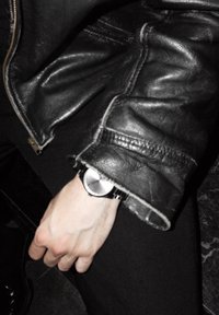 Hopeakylkinen kello, jossa on minimalistinen muotoilu ja musta hihna, kädessä, jonka päällä on nahkatakin hiha osittain peittämässä.