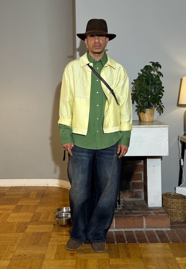 Mann trägt einen dunkelbraunen breitkrempigen Hut, gelbe Jacke, grünes Hemd mit Knöpfen, breite dunkle Jeans und braune Schuhe und steht drinnen in der Nähe eines Kamins und einer Pflanze.