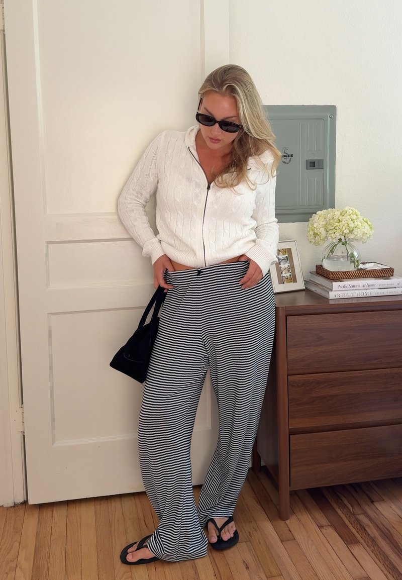 Femme portant des lunettes de soleil, un pull blanc zippé, un pantalon rayé et des tongs, se tenant à l'intérieur en tenant un sac noir près d'une commode en bois.