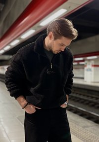 Juodas vilnonis megztinis su aukštu apykakle ir pusiau užtrauktuku, su išsiuvinėtais užrašais. Dėvimas su juodomis kelnėmis metro stotyje.