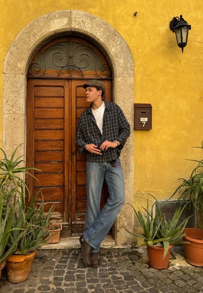Joven con gorra, camisa de cuadros y jeans apoyado en una puerta de madera arqueada, con suculentas en macetas y una pared amarilla con un buzón y una linterna.