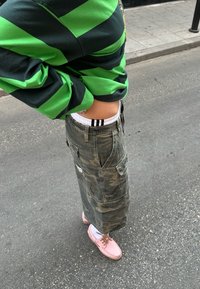 Grün und schwarz gestreiftes Langarmshirt; Tarnmuster Cargo-Rock; pinke Schuhe mit heller Sohle; weiße Socken mit schwarzen Akzenten.