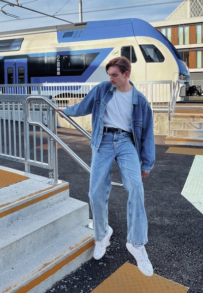 Chaqueta de mezclilla y jeans de tiro alto de color azul claro combinados con zapatillas blancas; de pie en escalones de concreto con una barandilla plateada, fondo de tren.