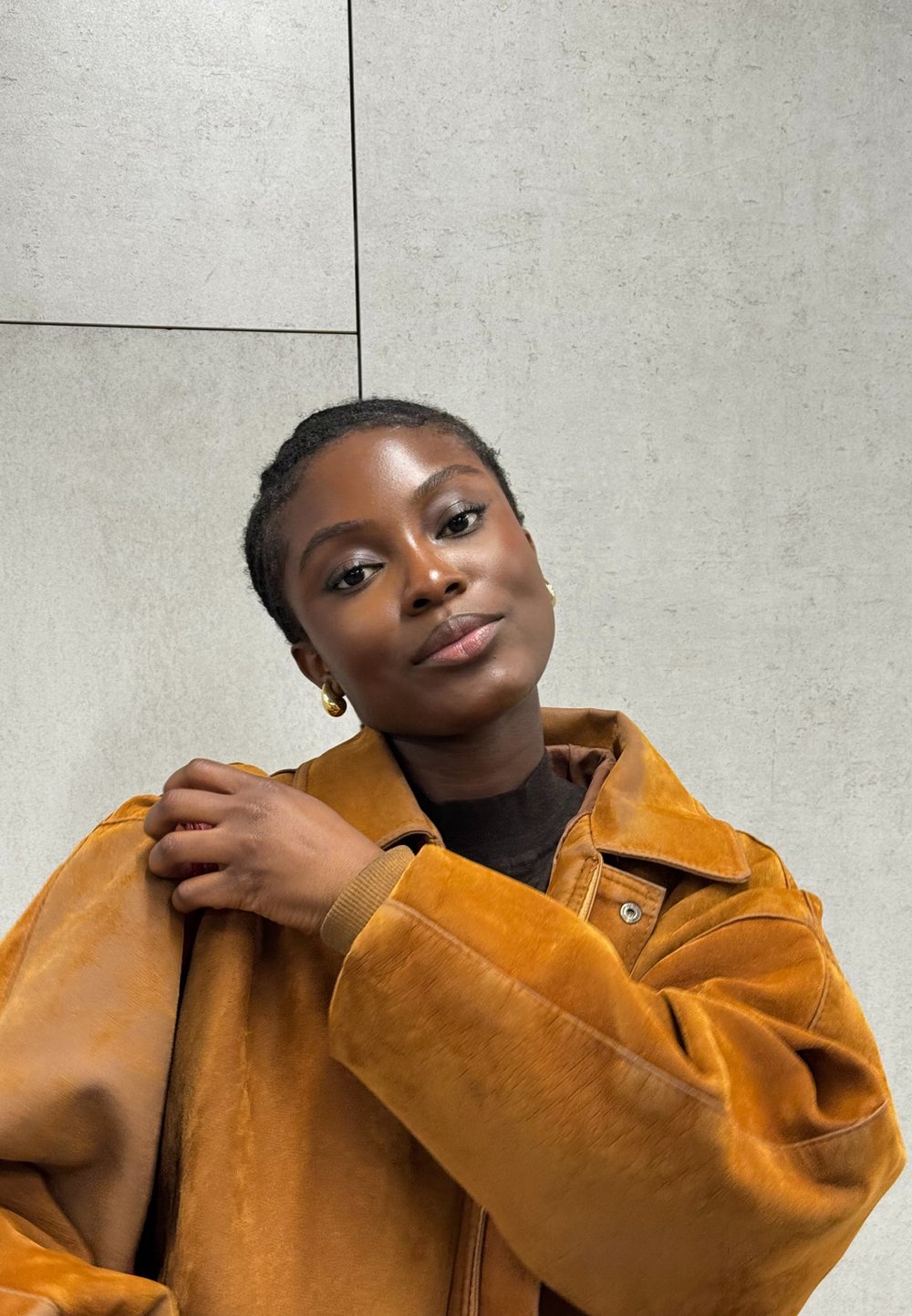 Veste oversized en daim marron avec un col et des coutures visibles. La personne qui porte la veste a de courtes tresses et des boucles d'oreilles en or, le tout sur un fond gris texturé.