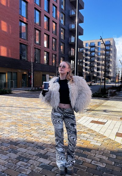 Joven con gafas de sol, chaqueta de peluche y pantalones estampados sosteniendo una taza de café, de pie en una calle empedrada junto a edificios modernos.